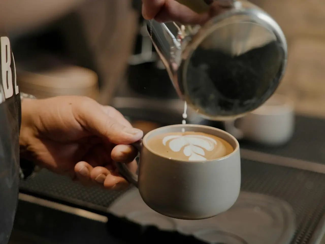 A barista pouring a latte with a rosetta in a grey cup