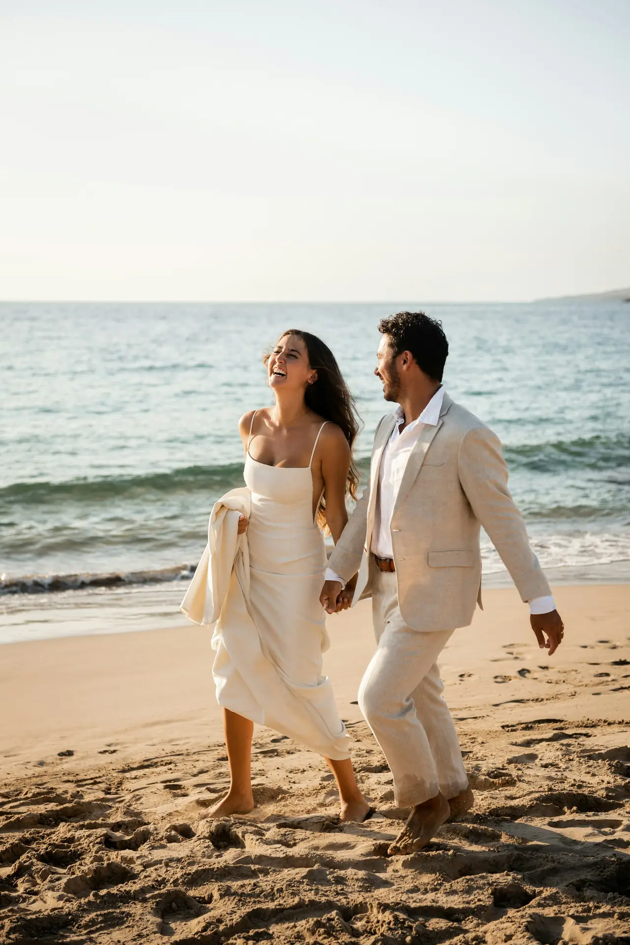 A couple in white walking hand in hand along the sand with the sea behind them