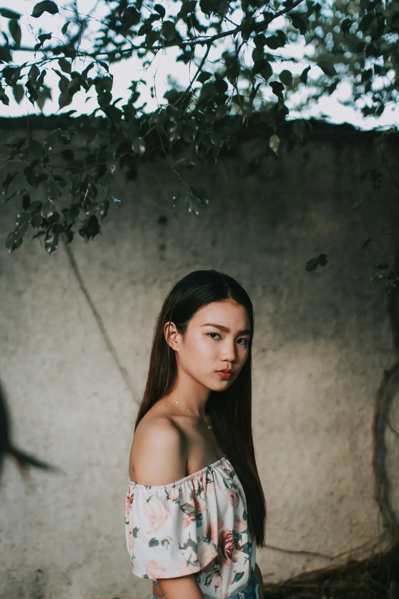 A woman in a floral off-the-shoulder top standing against a rough wall under leaves