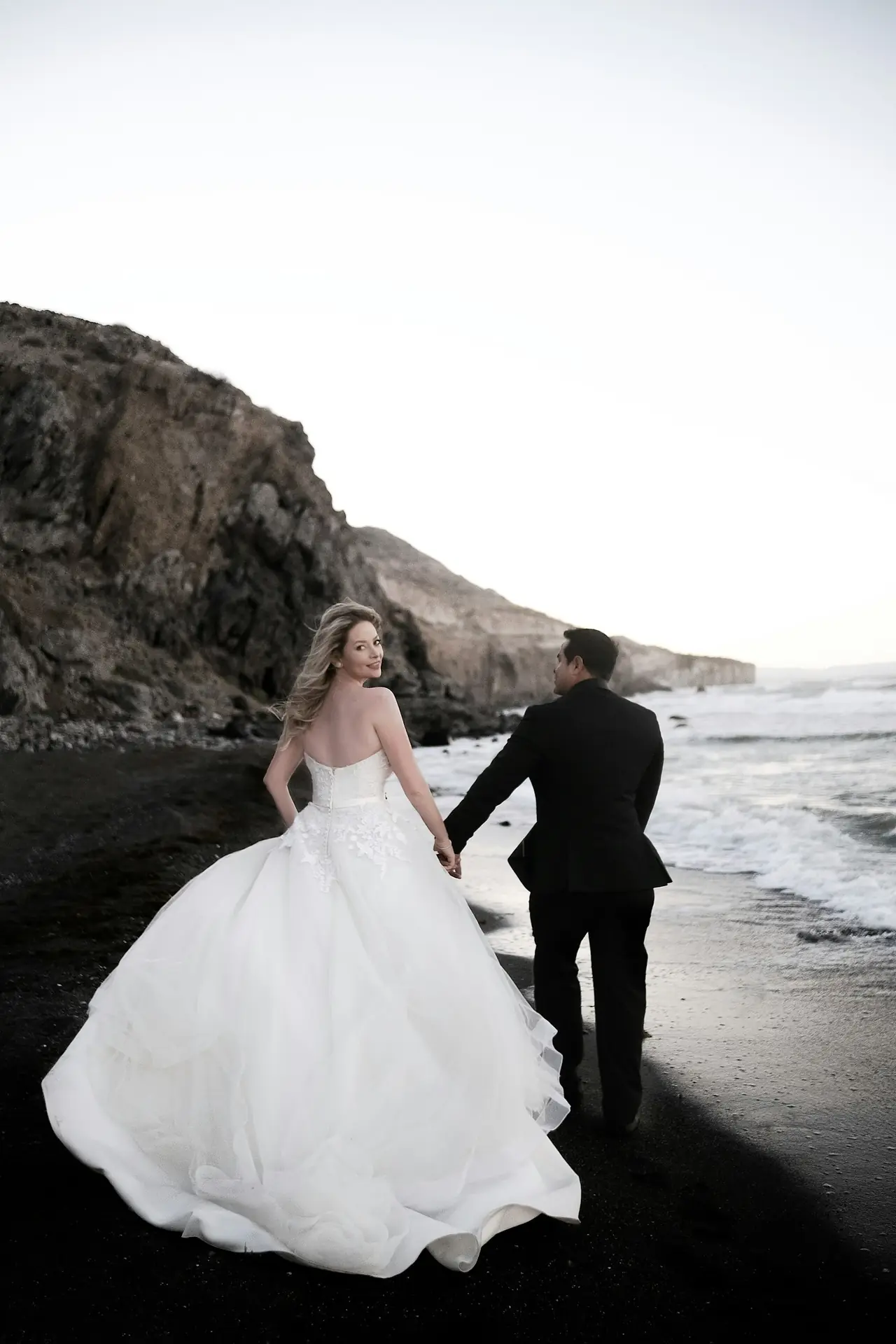 A bride in a full white dress walking with her groom along a dark beach below cliffs