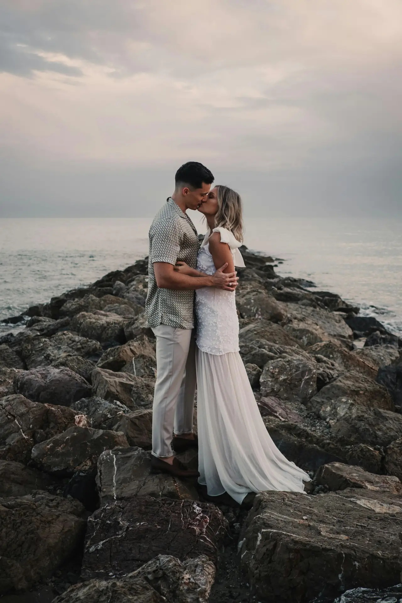 A couple embracing on a rocky breakwater under a pale sky