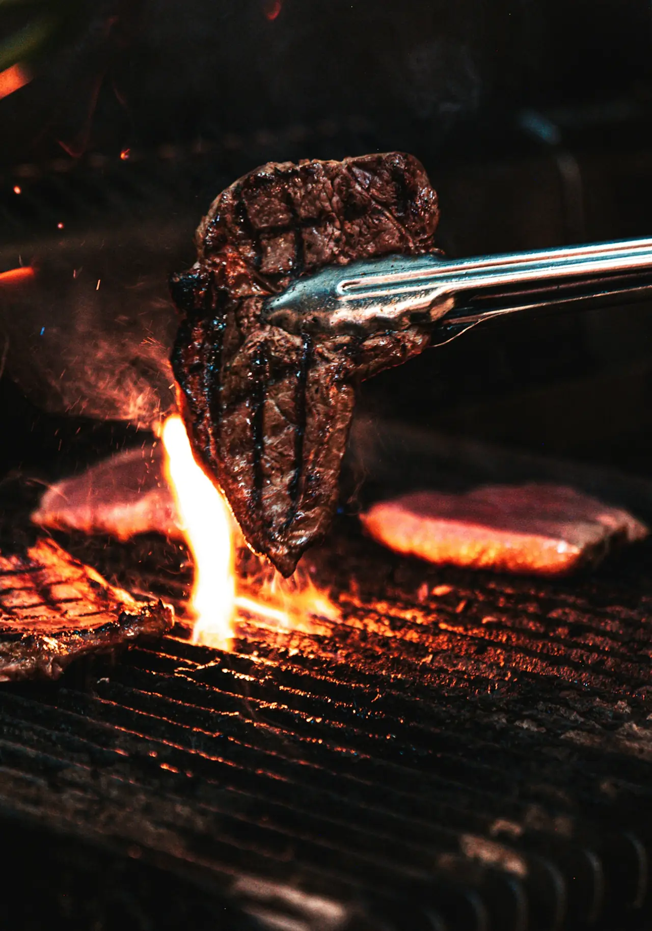 A steak lifted with tongs over a grill and open flame